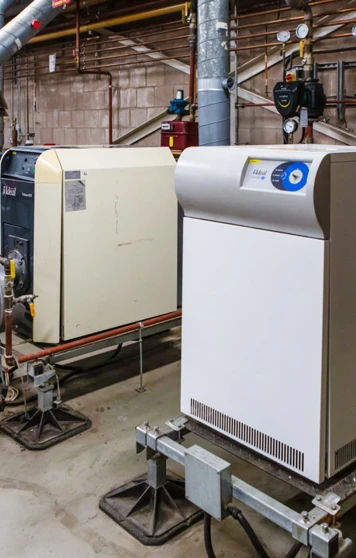 A row of industrial boilers in a building services training facility, connected to ventilation ducts and a complex piping system. A row of industrial boilers in a building services training facility, connected to ventilation ducts and a complex piping system.